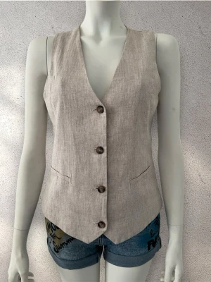 Beige linneväst med knappar - Stilren beige väst i linne med fyra bruna knappar framtill. Västen har v-ringad hals och en klassisk, något figurnära passform. Perfekt att styla över en t-shirt eller skjorta för en trendig look. #väst #linne