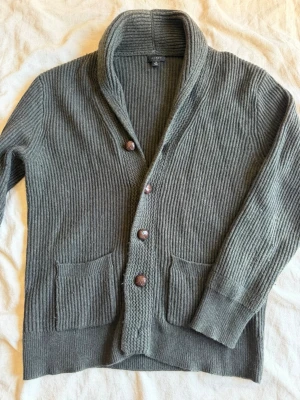 Grå kofta från J.Crew med sjalkrage - Mysig grå stickad kofta från J.Crew med sjalkrage och knäppning framtill. Koftan har två stora fickor och ribbstickad struktur som ger en avslappnad men stilren look. Perfekt för lager-på-lager och chill dagar.