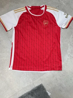 Arsenal matchtröja Saka #7 röd/vit - Snygg Arsenal matchtröja från Adidas i rött och vitt med guldiga detaljer på axlarna. Tröjan har klubbmärke på bröstet, Saka och nummer 7 på ryggen samt blixtmönster över hela framsidan. Perfekt för fotbollsfans!