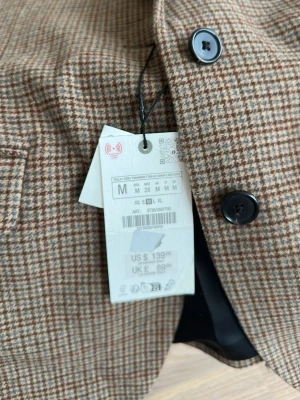 Brunrutig kavaj från Zara, M - Snygg brunrutig kavaj från Zara i storlek M. Klassisk modell med två knappar framtill, fickor med lock och helfodrad insida. Perfekt för en stilren och trendig look. Materialet är mjukt och har en vintageinspirerad känsla.