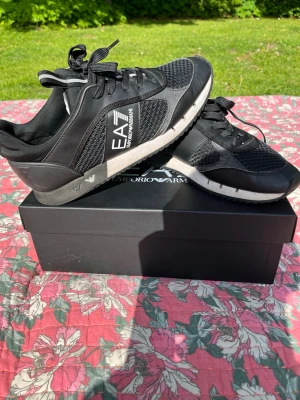 Svarta EA7 Emporio Armani sneakers - Snygga svarta sneakers från EA7 Emporio Armani med vit logga på sidan. Skorna har meshdetaljer för extra andningsförmåga och svarta snören. Sulan är svart och vit med diskreta logodetaljer. Perfekta för en trendig och sportig look.