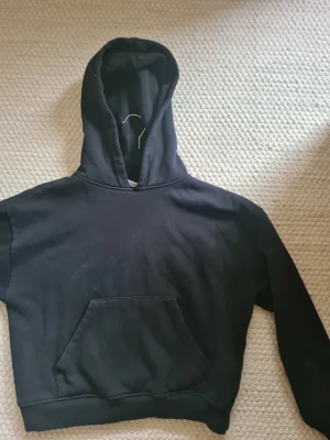 Svart hoodie med magficka - En klassisk svart hoodie med rymlig magficka. Hoodien har en avslappnad passform och ribbade muddar vid ärmslut och nederkant.