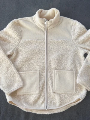 Krämvit teddyjacka från Pieces - Mysig teddyjacka i krämvitt från Pieces. Jackan har två stora fickor framtill, dragkedja och hög krage. Tillverkad i fluffigt teddy-material som ger en cool och avslappnad look. Perfekt för lager-på-lager-stil under kyliga dagar.