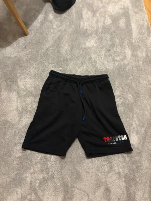 Svarta Trapstar shorts med snörning - Svarta shorts från Trapstar med elastisk midja och snörning. Tryckt Trapstar-logga i rött, vitt och grått på vänster ben. Perfekta för en avslappnad streetwear-look. Tillverkade i mjukt material för extra komfort.