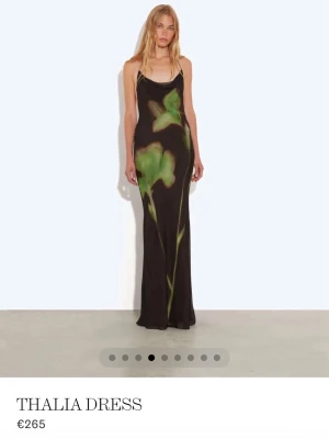 Svart långklänning Thalia Dress - Svart långklänning med tunna axelband och öppen rygg. Klänningen har ett unikt blommigt mönster i grönt och beige framtill. Snygg, figurnära passform och elegant siluett. Perfekt för dig som vill sticka ut med en cool och modern look.