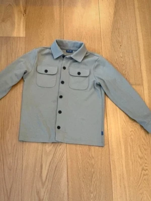 Only&sons ljusgrå overshirt - Stilren ljusgrå overshirt med klassisk krage och svarta knappar framtill. Två bröstfickor med lock och knappdetaljer ger en snygg look. Tillverkad i ett mjukt tyg som passar perfekt till lager-på-lager-stil.