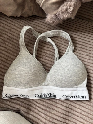 Grå sport-bh från Calvin Klein - Snygg grå sport-bh från Calvin Klein med vadderade kupor och brottarrygg. Resårbandet är vitt med svart logotyptryck runtom. Tillverkad i mjuk bomullsblandning för extra komfort.