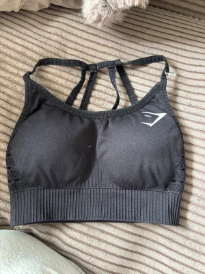 Svart sport-bh från Gymshark - Svart sport-bh från Gymshark med ribbad nederkant och flera smala axelband i ryggen. Bh:n har en diskret logga framtill och bak, samt meshdetaljer på sidorna för extra ventilation. Perfekt för träning och aktiva dagar.