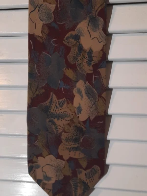 Mönstrad slips från Van Gils - Säljer en snygg slips från Van Gils i 100% siden. Slipsen har ett abstrakt blommönster i brunt, beige, blått och olivgrönt mot en vinröd bakgrund. Perfekt för att ge outfiten en unik touch.