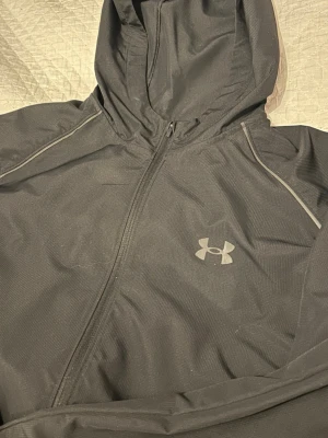 Svart vindjacka från Under Armour - Snygg svart vindjacka från Under Armour med huva och hel dragkedja framtill. Jackan har diskreta reflexdetaljer längs axlarna och Under Armour-logga på bröstet. Tillverkad i lätt och funktionellt material som passar perfekt för blåsiga dagar.