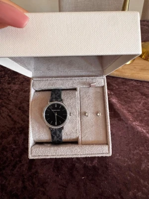 Svart klocka från Michael Kors - Stilren svart klocka från Michael Kors med silverfärgade detaljer. Klockan har ett svart band med mönster och en ram med små glittrande stenar runt urtavlan. Perfekt accessoar för att lyfta din outfit.