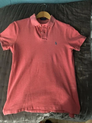 Rosa pikétröja från Ralph Lauren - Klassisk rosa pikétröja från Ralph Lauren med korta ärmar och krage. Tröjan har knappar framtill och den ikoniska broderade loggan på bröstet. Perfekt för en avslappnad men ändå stilren look.