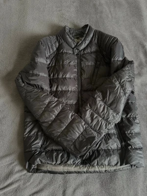 Svart dunjacka med ståkrage - Snygg svart dunjacka med ståkrage och dragkedja framtill. Jackan har quiltad design med horisontella sömmar och en diskret bröstficka med dragkedja. Perfekt för kalla vinterdagar och enkel att matcha med olika outfits.