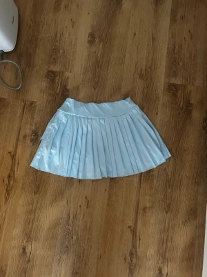 Ljusblå plisserad kortkjol - Snygg ljusblå kortkjol med plisserad design. Kjolen har en bred linning och ett glansigt tyg som ger en fräsch och trendig look. Perfekt för dig som vill sticka ut med en stilren och ungdomlig vibe.