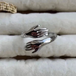Silvrig ring med handmotiv - Unik silvrig ring i med två händer som möts framtill. Ringen har en öppen design och blanka ytor som ger en modern och cool look. Perfekt för dig som gillar statement-smycken och vill ha något annorlunda.