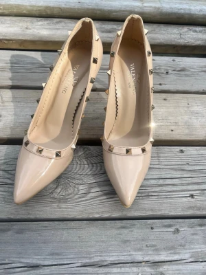 Beige lackpumps med nitar - Snygga beige pumps från Valentino i lackat material med spetsig tå och höga klackar. Dekorerade med coola guldfärgade nitar längs kanten för en edgy look. Perfekta för dig som vill sticka ut lite extra.