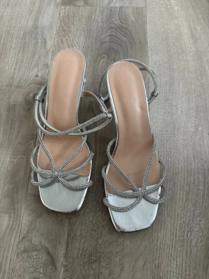 Silvriga pumps med strassdetaljer - Säljer ett par eleganta silvriga pumps med tunna remmar dekorerade med strass. Skorna har fyrkantig tå och justerbar ankelrem för extra komfort. Perfekta för dig som vill sticka ut med en glittrig och trendig look.