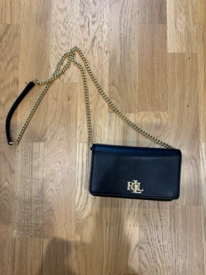 RL Svart kuvertväska med guldkedja - Stilren svart kuvertväska från Ralph Lauren med elegant guldfärgad RL-logga framtill.  Lyxig!! Men används dessvärre aldrig….