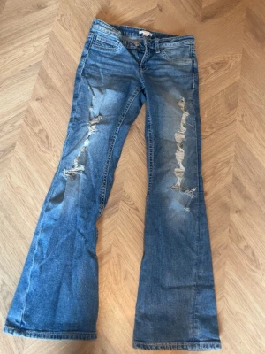 Blå bootcut jeans med hål - Säljer ett par ljusblå bootcut jeans med slitningar på båda benen och snygga detaljerade bakfickor. Jeansen har låg midja och klassisk femficksdesign. Perfekta för en avslappnad och trendig look.
