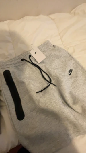 Nike tech shorts gråa - Snygga gråa joggers från Nike med svart dragsko i midjan och en svart dragkedjeficka på sidan. Byxorna har Nike-logga broderad på benet och är tillverkade i mjukt material, perfekta för träning eller chill.