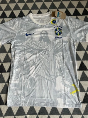 Vit Brasilien Nike fotbollströja M - Säljer en vit fotbollströja från Nike med Brasilien-emblem och blå Nike-logga. Tröjan har ett detaljerat grått mönster med Kristusstatyn och andra brasilianska motiv. Rund hals och korta ärmar. Perfekt för dig som älskar fotboll och snygg design.