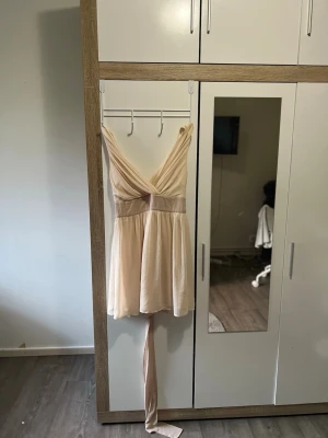 Kort ljus klänning med omlott och knyt - Söt kort klänning i ljus beige ton med omlottliv och djup v-ringning. Klänningen har bred midja och långa knytband som kan knytas i midjan och har även en liten dragkedja där bak. Tillverkad i ett mjukt, lätt material som ger ett fint fall.