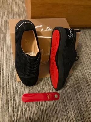 Svarta sneakers från Christian Louboutin - Stilrena svarta sneakers från Christian Louboutin i mocka med klassisk röd sula och svart snörning. Låg modell med diskret logga på hälen och insidan. Perfekta för dig som vill ha en lyxig och unik touch på din outfit.
