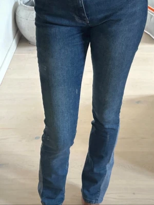 Blå bootcut jeans med hög midja - Säljer ett par klassiska blå bootcut jeans med hög midja och raka ben. Jeansen har en snygg tvätt och är tillverkade i ett mjukt denimtyg som sitter skönt. Perfekta till sneakers eller boots för en avslappnad look.
