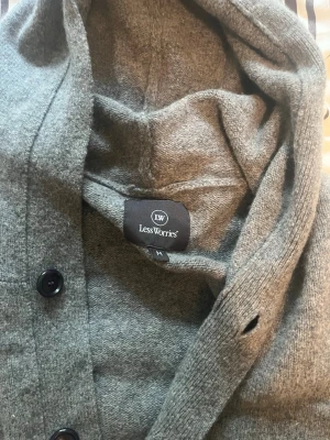 Grå hoodiekofta från Less Worries - Mysig grå kofta med huva från Less Worries. Koftan har knäppning framtill med svarta knappar, två fickor och ribbade muddar. Perfekt för lager på lager och chill dagar. Materialet känns mjukt och bekvämt.