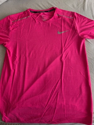 Nike träningsset rosa & svart - Snyggt träningsset från Nike med en rosa t-shirt i funktionsmaterial och svarta shorts med dragsko och fickor. Båda plaggen har Nike-logga i silver. Perfekt för löpning eller gym. T-shirten har rund hals och kort ärm, shortsen är lätta och luftiga.