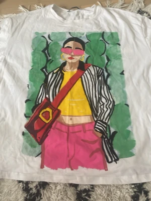 Vit t-shirt med grafiskt tryck från Zara - Vit t-shirt från Zara med ett stort grafiskt tryck framtill som föreställer en person i färgglada kläder och solglasögon. T-shirten har rund hals och korta ärmar. Perfekt för dig som gillar statement-plagg och konstnärliga motiv.