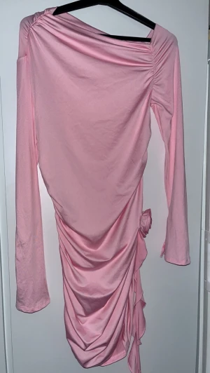Rosa långärmad omlottklänning - Säljer en rosa långärmad klänning med omlottdetalj och drapering längs sidan. Klänningen har en dekorativ rosett i midjan och är tillverkad i ett mjukt, stretchigt material. Perfekt för dig som gillar en feminin och trendig stil. Perfekt för fest !