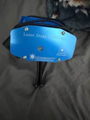 Blå Laser Stage Lighting enhet - Laser Stage Lighting i metall med blå färg, kompakt design och flera inställningsmöjligheter. Utrustad med strömkabel och stativ, perfekt för att skapa ljuseffekter på fester eller events. Klass IIIA laserprodukt med ventilationsgaller och kontrollpanel på baksidan.