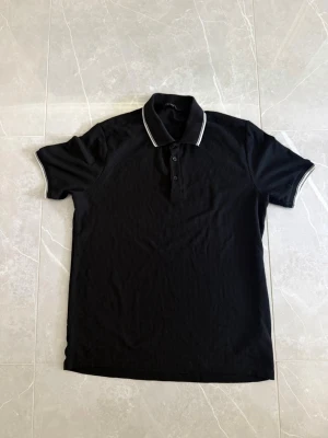 Svart pikétröja med vit kant - Klassisk svart pikétröja med vit kant på kragen och ärmsluten. Tröjan har knappar framtill och är kortärmad. Perfekt till en avslappnad men ändå stilren look.