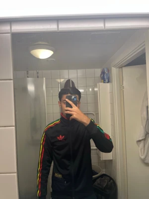 Svart Adidas track jacket XS - Säljer en svart Adidas track jacket med färgglada ränder i rött, gult och grönt längs ärmarna. Tröjan har dragkedja framtill, hög krage och Adidas logga broderad på bröstet. Perfekt för en sportig och retro look.