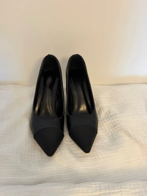 Svarta pumps med pärldetalj - Snygga svarta pumps med spetsig tå och unik pärldetalj på klacken. Skorna har en klassisk siluett och är tillverkade i ett slätt material som ser ut som konstläder. Perfekta för dig som vill ha en stilren men ändå trendig look. Pärl designen syns inte på sista bilden då de inte är riktiga cc klackar