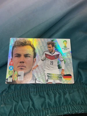 Glittrig fotbollssamlarbild Mario Götze - Säljer en glittrig samlarbild från FIFA World Cup Brasil med Mario Götze i tysk landslagströja. Kortet har holografisk yta, färgglada detaljer och visar både porträtt och actionbild. Perfekt för dig som gillar fotboll och samlarbilder.