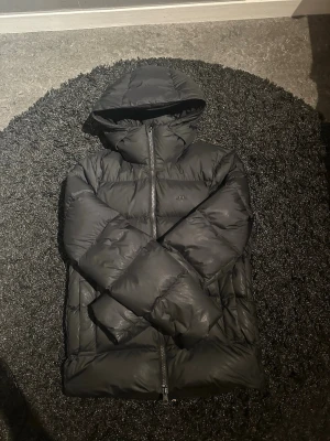 Svart pufferjacka med huva - Säljer en svart pufferjacka med huva och dragkedja framtill. Jackan har två sidofickor och en quiltad design som ger en riktigt puffig look. Perfekt för kalla vinterdagar och riktigt skön att bära. Materialet är glansigt och vattenavvisande.