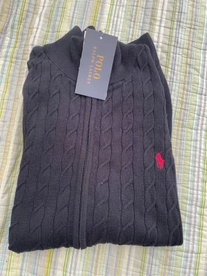 Svart kabelstickad kofta Polo Ralph Lauren - Kabelstickad svart kofta från Polo Ralph Lauren med dragkedja framtill och röd broderad logga på bröstet. Hög krage och klassisk design som passar till det mesta. Perfekt för dig som gillar stilrena och tidlösa plagg.