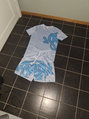Syna world x central cee - Säljer ett par ljusblå shorts med stort blått graffiti-inspirerat tryck. Shortsen har resår i midjan och matchar perfekt med t-shirten i samma stil. Materialet känns som polyester och passar bra till sommaren.