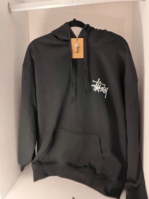 Svart hoodie från Stussy med logga - Svart hoodie från Stussy med vit logga tryckt på bröstet. Klassisk känguruficka framtill och justerbar huva med snören. Tillverkad i mjukt material som känns skönt mot huden. Perfekt för en avslappnad streetwear-look.