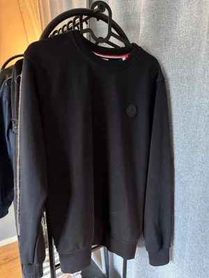 Svart sweatshirt från Moncler - Stilren svart sweatshirt från Moncler med rund halsringning och diskret Moncler-logga på bröstet. Tröjan har ribbade muddar vid ärmslut och nederkant samt en snygg detalj med röd, vit och blå rand på insidan av kragen.