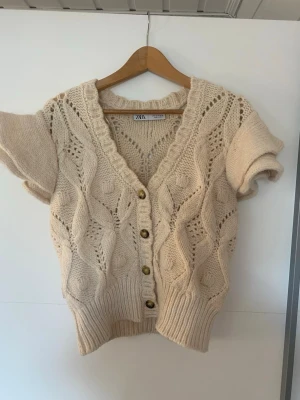 Beige kortärmad stickad kofta Zara - Supersöt beige stickad kofta från Zara med korta ärmar och djup V-ringning. Koftan har dekorativa träknappar framtill och ett fint hålmönster i garnet. Ribbstickad kant nedtill och vid ärmslut. Perfekt att slänga över en topp för en mysig look.
