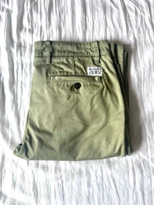 Olivgröna chinos från Norse Projects  - Snygga olivgröna chinos från Norse Projects  med klassisk rak passform. Byxorna har sidofickor, bakficka med knapp och bälteshällor. Tillverkade i mjukt bomullstyg som ger en skön känsla och stilren look.