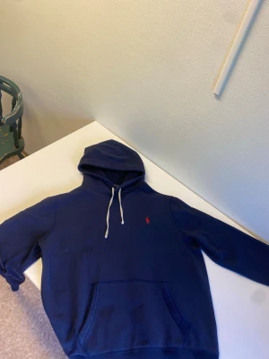 Mörkblå hoodie från Polo Ralph Lauren - Säljer en mörkblå hoodie från Polo Ralph Lauren med klassisk huva och vita dragsnören. Tröjan har en känguruficka framtill och den ikoniska röda Polo-loggan broderad på bröstet. Perfekt för en chill och stilren look.