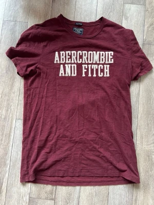 Vinröd Abercrombie & Fitch t-shirt - Snygg vinröd t-shirt från Abercrombie & Fitch med klassisk vit logga tryckt framtill. T-shirten har rund hals och korta ärmar, perfekt för en avslappnad och trendig look. Tillverkad i mjuk bomull för skön komfort.