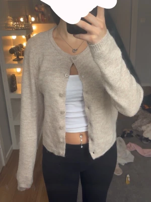 Beige knappad kofta  - Mysig beige kofta med rund halsringning och knappar framtill. Långärmad modell i mjuk ullblandning som passar perfekt till lager på lager. Enkel och stilren design som är lätt att matcha med olika outfits.
