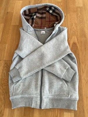 Grå zip hoodie från Burberry helt ny  - Säljer en grå zip hoodie från Burberry med klassiskt rutigt foder i huvan. Hoodien har dragkedja framtill, två fickor och ribbade muddar vid ärmar och nederkant. Perfekt för dig som gillar stilrena plagg med exklusiva detaljer.