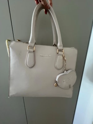 Beige handväska med gulddetaljer - Stilren beige handväska från Michael Shannon med två rejäla handtag och guldfärgade metalldetaljer. Väskan har en praktisk dragkedja baktill och ett matchande litet myntfodral som hänger framtill. Tillverkad i slätt konstläder med enkel, fyrkantig form.
