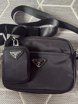 Svart axelväska  - Snygg svart axelväska   nylon med justerbar axelrem och silverfärgade detaljer. Väskan har en extra liten avtagbar ficka framtill och klassiska-loggor i metall. Perfekt för dig som vill ha en stilren och trendig accessoar.